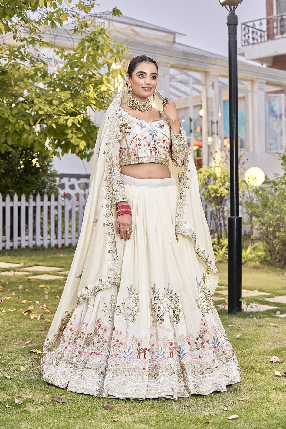 Woman in a traditional white embroidered outfit standing outdoors with greenery and a building in the background.